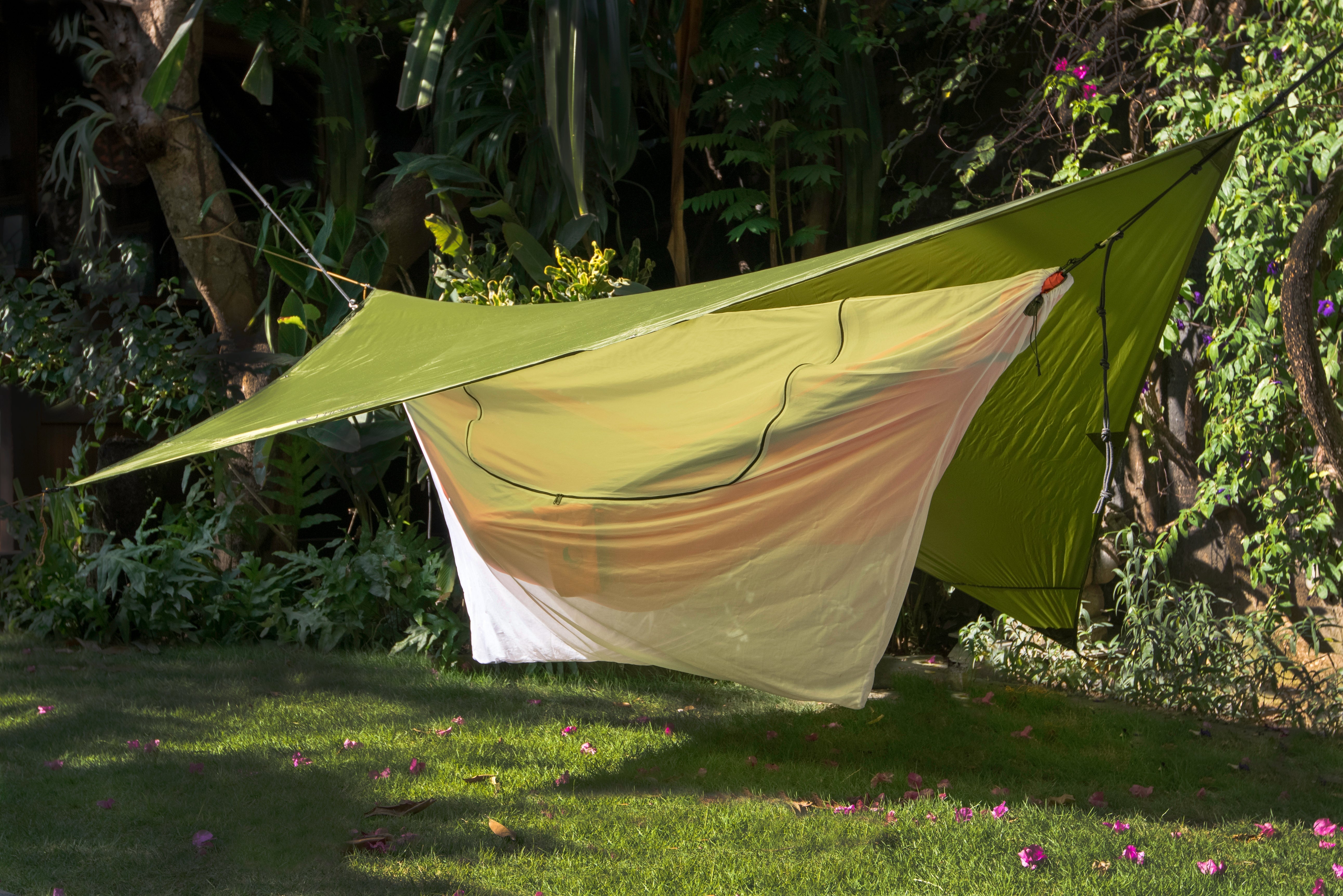 Weiße 360° Mückennetz-Hülle über einer Hängematte mit grünem Tarp als Regenschutz, gespannt im tropischen Garten mit Sonnenlicht und Schatten