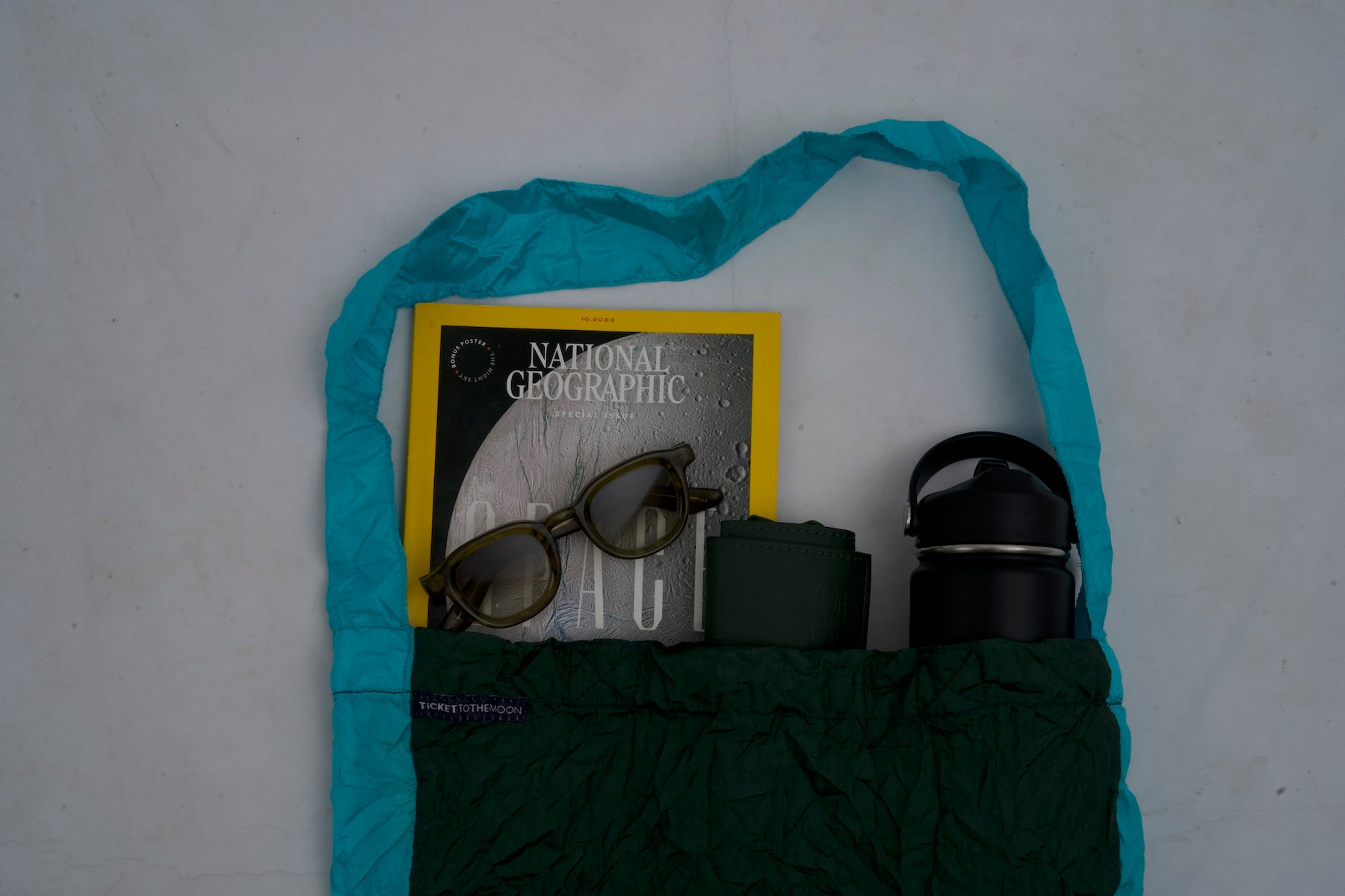 Kleine ECO BAG von Ticket to the Moon in Dunkelgrün und Türkis, gefüllt mit einer National Geographic Zeitschrift, Sonnenbrille, Geldbörse und Trinkflasche, flachliegend auf hellem Hintergrund.