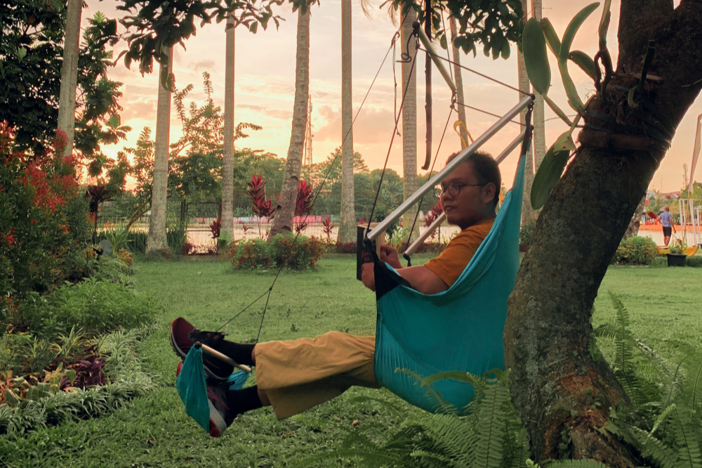 Person lehnt entspannt im türkisfarbenen MOON CHAIR mit Fußstütze an einem Baum, im Hintergrund tropischer Garten bei Sonnenuntergang.