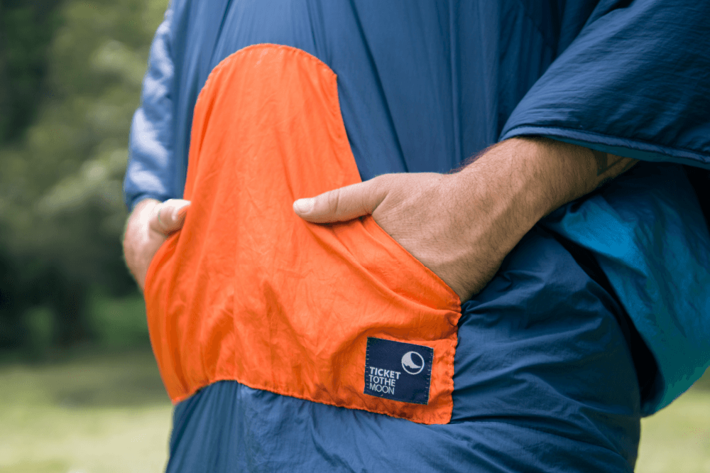 Nahaufnahme der großen orangefarbenen Vordertasche der Moonblanket mit beiden Händen darin, gut sichtbar ist das Ticket to the Moon Logo auf der Tasche