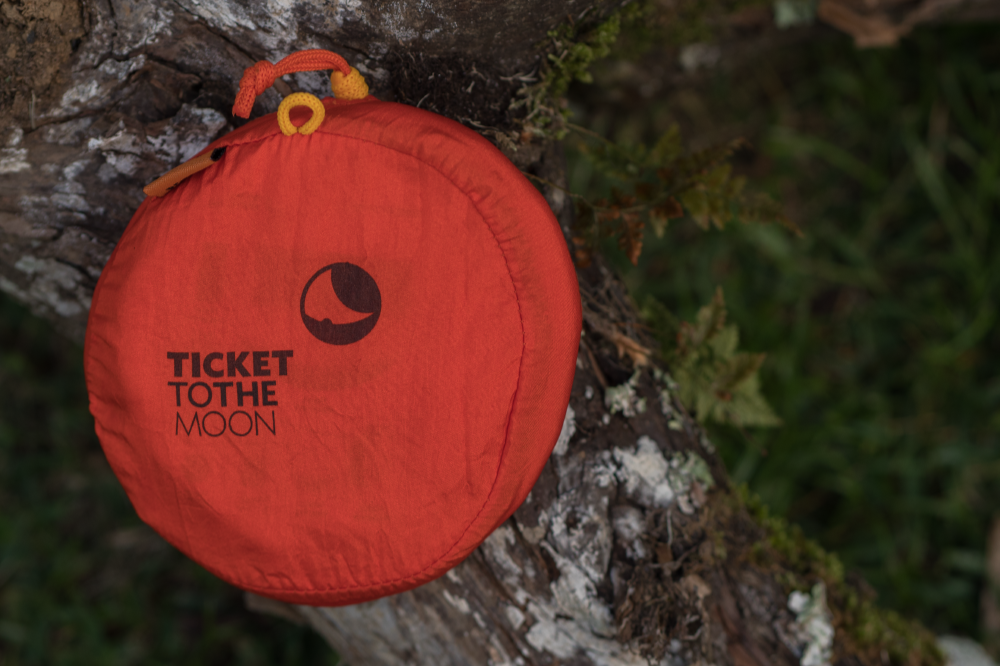 Rote Moondisc mit „Ticket to the Moon“-Logo liegt auf einem moosbedeckten Baumstamm im Grünen.