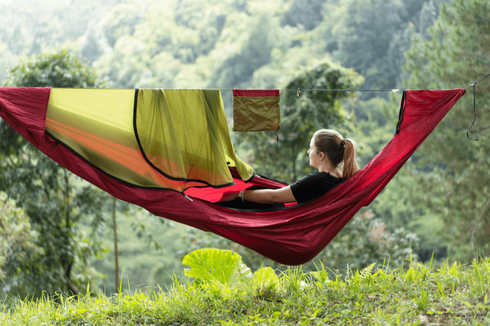 PRO HAMMOCK in Rot mit integriertem gelb-grünem Moskitonetz, aufgehängt am Waldrand mit Ausblick ins grüne Tal, eine Frau liegt entspannt darin.