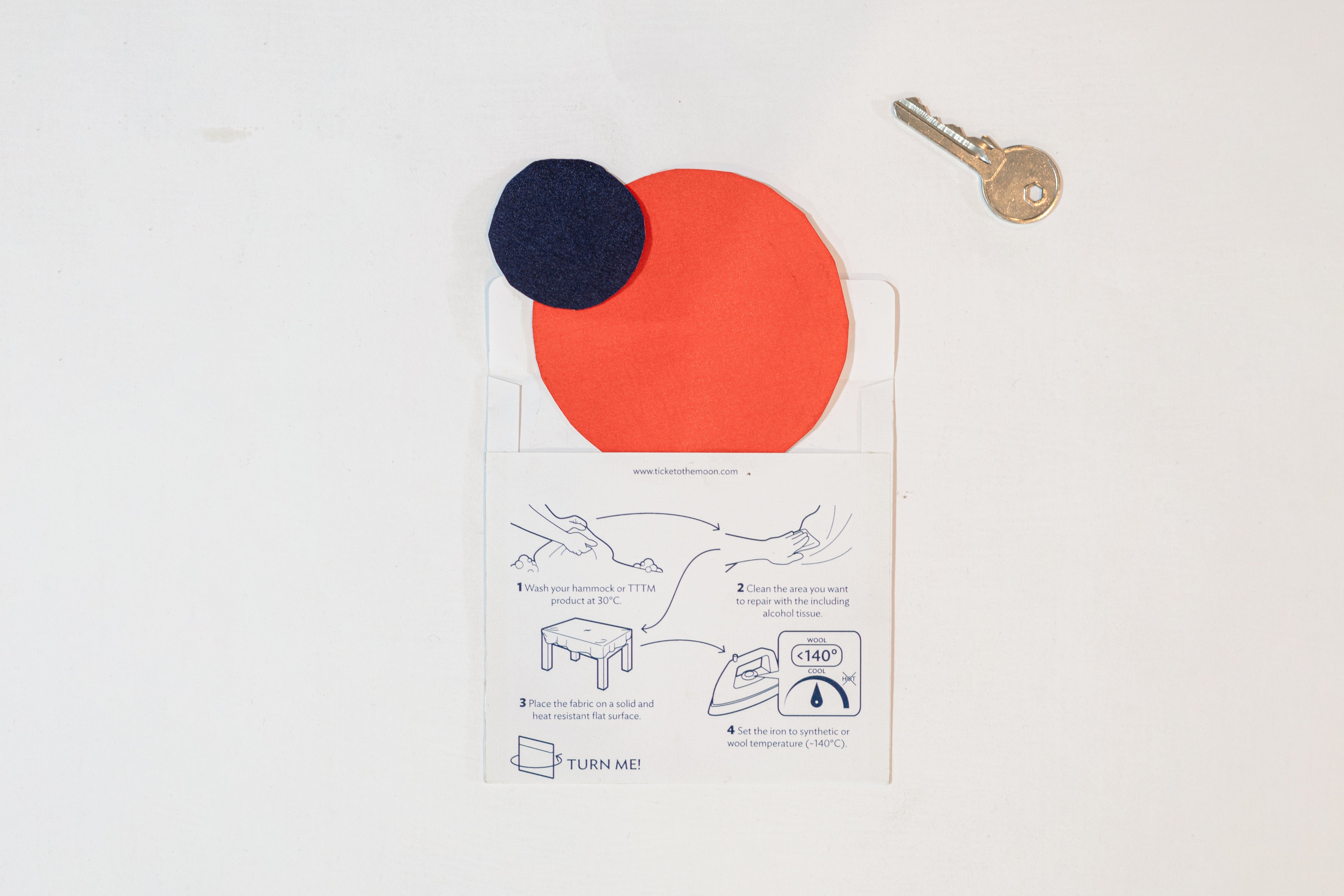 Reparaturset mit zwei runden Nylon-Patches in Dunkelblau und Orange in unterschiedlichen Größen, beiliegender Anleitung mit Bügelschritten und Schlüssel als Größenvergleich – geeignet für Hängematten- oder Stoffreparaturen.