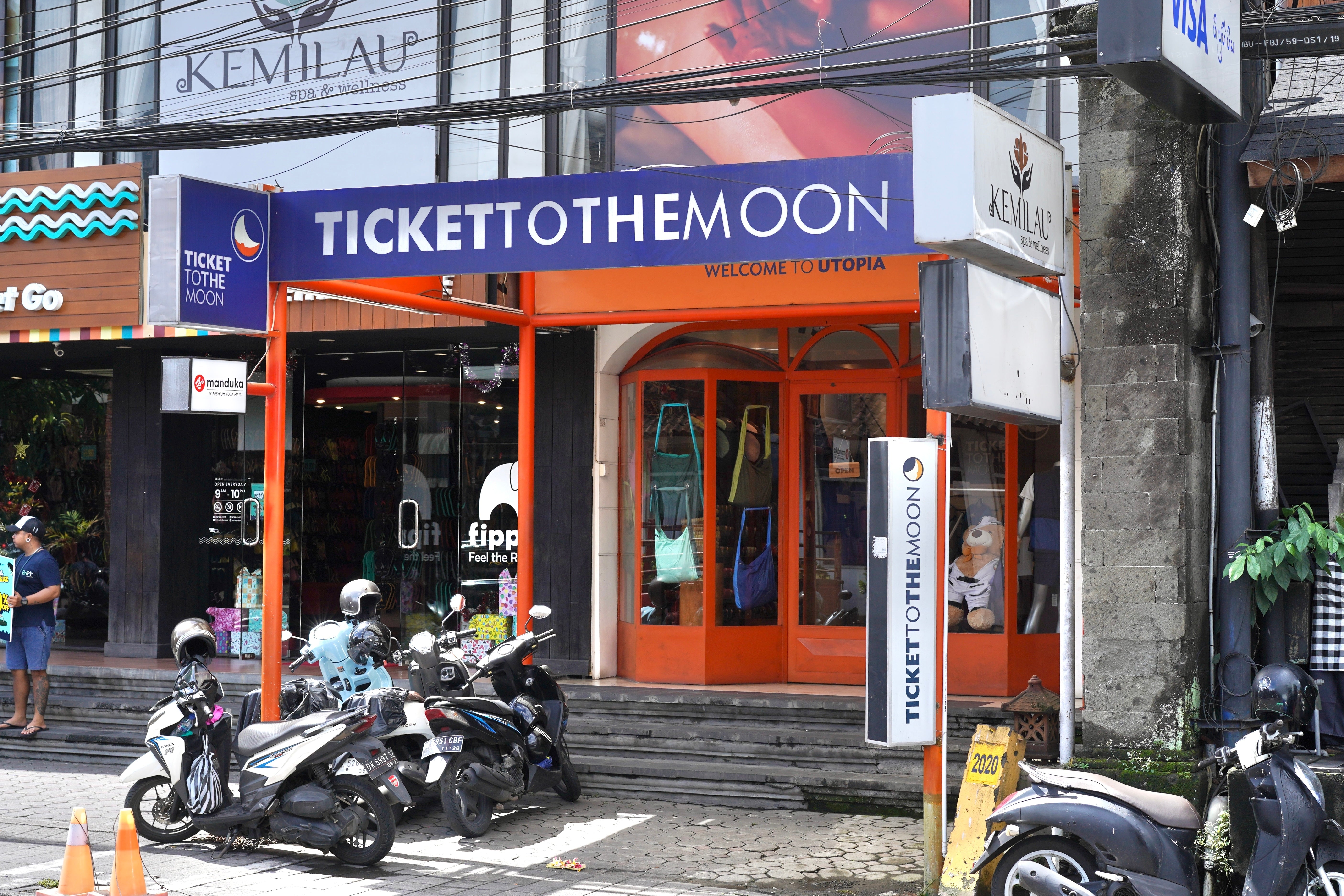 Straßenansicht eines Ticket to the Moon Stores in Bali/Ubud mit orangefarbenem Eingang, Rollern davor und farbenfroher Schaufensterdekoration mit Taschen und Accessoires.