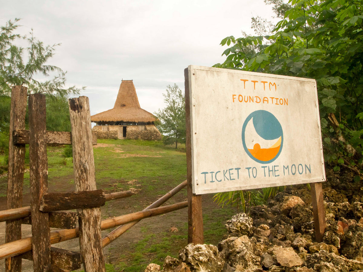 Holzschild der TTTM Foundation mit Logo vor einem traditionellen Haus mit strohgedecktem Dach auf Sumba, Indonesien